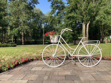 Artistic 3D white bicycle sculpture in outdoor park setting, surrounded by trimmed grass and red flowers, with lush green trees and clear blue sky in the background Real Kyiv, Ukraine 06-02-2024 clipart