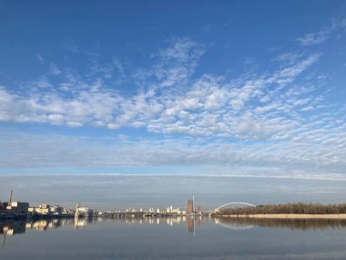 Suyun yansıttığı sakin bir açık hava sahnesi, uzak mesafelerde kentsel ufuk çizgisi, dağınık bulutlar ve Ukrayna, Kyiv, 11-02-2024 şehir manzarası yakınlarındaki olası park veya eğlence alanı.
