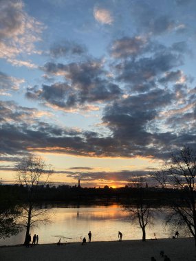 Durgun bir göl ya da nehir üzerinde sakin bir günbatımı sahnesi. İnsanlar kıyılarda geziniyor. Şehir silueti uzaklardan görünüyor.