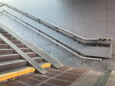 Gri basamaklı iç merdiven, metalik korkuluk, sarı güvenlik işareti, yan duvarda olası su hasarı, gri tavan kiremitleri veya paneller, kamu alanlarını düşündüren faydacı atmosfer