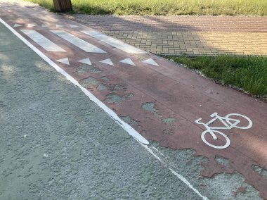 Görüntü, kaldırımda işaretli bir bisiklet şeridi ve çift yönlü kullanım için işaretli oklar göstermektedir. Yüzey asfalttır, bazı yerlerde giyilmiştir ve arka planda çimenler ve bir elektrik direği vardır.