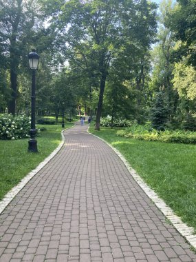 Yeşillik ve yapraklı ağaçların arasında kıvrılan kırmızı tuğlalı yolu olan sakin bir park, yol boyunca dinlenme yerleri sunan uzun bir sokak lambası, kentsel yetenek ekler ve dista 'da binalar görülebilir.