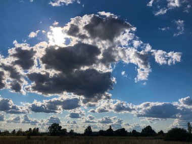 Image of a bright day sky with large, fluffy white clouds, one obscuring the sun for an intriguing play of light and shadow Distant landscape of green fields under a blue sky with scattered white cl clipart