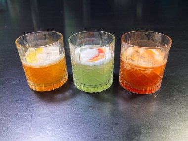 Image of three drinks, each presented in a clear glass on a reflective surface Each drink has a distinct appearance cloudy yellow-green fruity or herbal, murky light brown possibly honey, cream, clipart