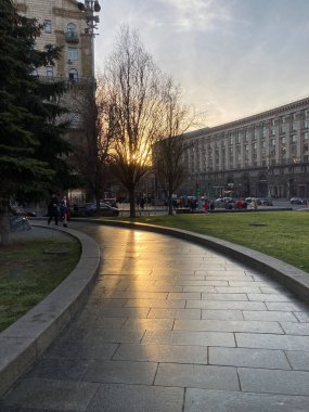 Tranquil urban scene at dawndusk, warm orange and blue sky, European buildings with green roofs, cobblestone path leading to viewer, recent rainfall, bare trees in background, late autumn or winter, clipart