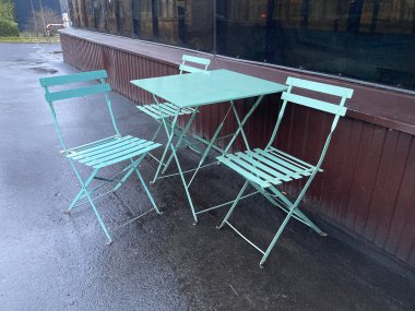 A compact set of green outdoor furniture consisting of two folding chairs and a round table on a potentially moist surface, under overcast daylight with blurred background elements clipart