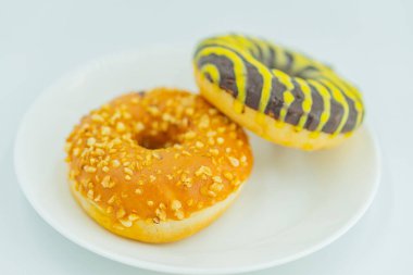 Açık gri arka planda beyaz bir tabakta iki donut ön planda yuvarlak, jöleli ve fındık kaplı muhtemelen badem ya da fıstık, arkada ise şerit var.