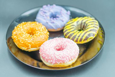 Beş renkli donut koyu bir tabakta sıralanır, her birinin üzerinde eşsiz bir krema ve tepsi vardır. Çörekler arasında açık kahverengi ve turuncu çekirdekli bir tane, mor kremalı ve parıltılı bir çörek vardır.