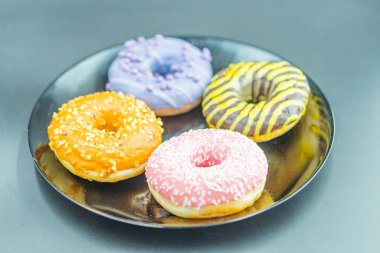 Image showcases a neat arrangement of 4 colorful donuts with various toppings on a black plate Toppings include glaze with sprinkles, solid purple icing, yellow drizzle, and pink confetti sprinkles clipart