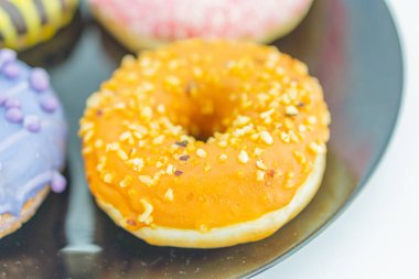 Close-up of three donuts on a reflective surface, with the central one glossy orange and nutty, and others partially obscured The image has a shallow depth of field, bright lighting, and a plain whi clipart