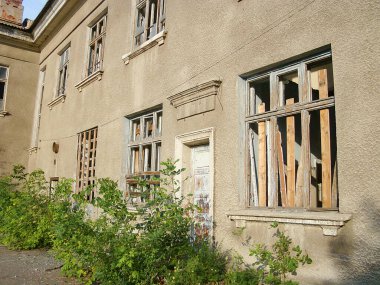 Terk edilmiş, çok katlı, bozulma işaretleri olan bir bina, tahtalanmış hasarlı pencereler, bitkisel büyüme, soyulmuş boya, pas lekeleri, yerde yosun, belirgin bir iz ya da konum yok.