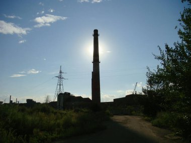 Görüntü, önde büyük bir bacası olan terk edilmiş bir sanayi sahnesini gösteriyor. Düz olmayan bitkiler, paslanmış metal yapılar, döküntüler, ağaçların siluetleri, mavi gökyüzü, güneş ışığı akışı.