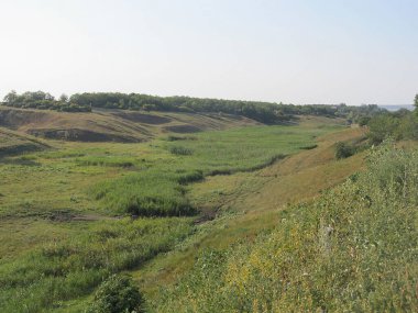 Yeşil tepeleri yuvarlayan doğal bir manzara, bir su yolu, ve el değmemiş yeşil bir gökyüzü, görünür bir yapı yok.