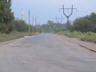 Bulutlu bir gökyüzü altında kırsal ya da yarı kırsal alanda elektrik direkleri, elektrik hatları, ağaçlar ve ekipman konteynırları olan yıpranmış asfalt bir yol. Araç ya da yayalar görünmüyor.