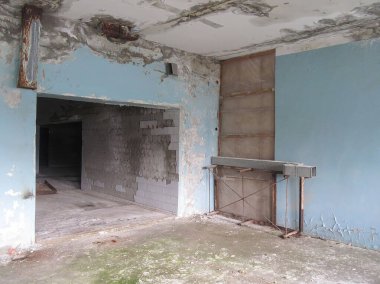 Görüntü harap olmuş mavi duvarlı bir iç mekan, karanlığa açılan açık bir kapı ve metal bir yapı gösteriyor.