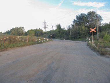 Görüntü, toprak bir yol, lastik izleri, çimenlik alan, dağınık çalılar ve yaprak döken ağaçlar, bulutlu gökyüzü, elektrik direkleri, tren yolu kesişen bir yolda demiryolu uyarı levhası, Candi
