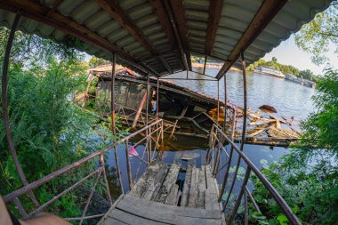 Kısmen batık, bitkilerle harap olmuş bir ahşap iskele, üzerinde duran birinin perspektifinden çekilmiş, su, ağaçlar ve uzakta küçük bir tekne ile çevrili.
