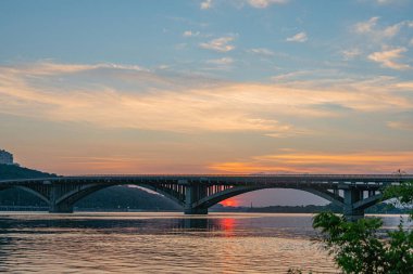 Gün batımı ya da gündoğumu sahnesinde büyük, çok kemerli bir köprü ile bir nehir gölü üzerinde gökyüzü pembemsi turuncudan açık maviye geçiş yapar.