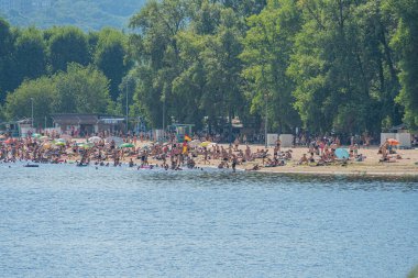 Açık bir günde plaj sahnesi güneşlenme, yüzme ve dalgıç şemsiyeleri gibi çeşitli aktivitelerle gölgeli bir çevre sunar. Doğal olarak ağaçlar Ukrayna, Kyiv, 06-17-2024 'ün arkasında sıralanır.