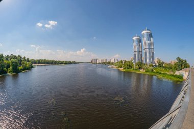 Görüntü, her iki tarafında modern yüksek binaların bulunduğu sakin bir nehir manzarasını gösteriyor. Sol kıyıda üç önemli yapı yer alıyor: Bir basamak gökdelen, bir gl Kyiv, Ukrayna 06-17-2024