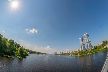 Görüntü, yüksek binaları, yansıtıcı cam cepheleri ve belirgin yükseklikteki kulesi ile çağdaş bir kentsel rıhtımı tasvir ediyor. Bölge, yapıyı yansıtan sakin sulara sahip Kyiv, Ukrayna 06-17-2024