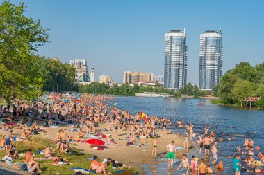Şehir ve doğal elementlerin karışımından oluşan canlı nehir kenarı sahnesi. İnsanlar suda güneşleniyor, yüzüyor ve eğleniyor. Arka planda iki gökdelenmiş bina, Kyiv, Ukrayna 06-17-2024 'ü tanımlıyor.
