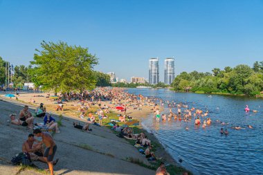 Canlı bir şehir plajını modern yüksek binaların arka planına karşı canlı bir görüntü sergiliyor. İnsanlar kumlu sahilde su aktiviteleriyle rahatlarken, yüksek yapılar Kyiv, Ukrayna 06-17-2024