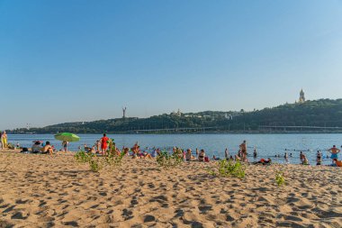 Huzurlu bir sahil sahnesinde doğa ve mimarinin bir karışımı sergileniyor, kumlu sahilde insanlar, yemyeşil, büyük bir köprü ve uzak görkemli bir bina Kyiv, Ukrayna 06-17-2024