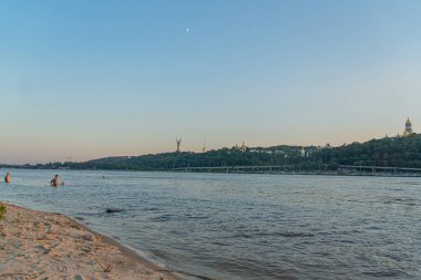 Tranquil riverside scene, dusk or dawn Bridge with multiple arches spanning river Traditional European style City skyline partially obscured by nature Urban development, Kyiv, Ukraine 06-17-2024 clipart