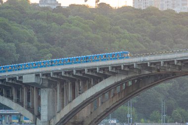 Image showcases a vibrant train moving across an elevated railway line over water, supported by a bridge with arches The scene is part of a cityscape where architecture harm Kyiv, Ukraine 06-17-2024 clipart