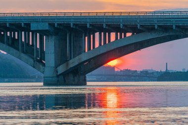 Günbatımına karşı kurulmuş bir asma köprü, doğanın ortasında yakalanır. Endüstriyel yapı su üzerinde uzanır ve güç için işlevsel tasarımını gösterir. Du Kyiv, Ukrayna 06-17-2024