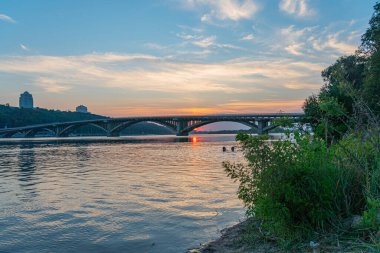 Güneşin doğuşunda ya da batışında sakin bir rıhtım sahnesi, nehrin üzerinde modern, kemerli bir köprü, sakin suları yansıtıyor, köprü yeşil alanları birleştiriyor ve Kyiv, Ukrayna, 06-17-2024