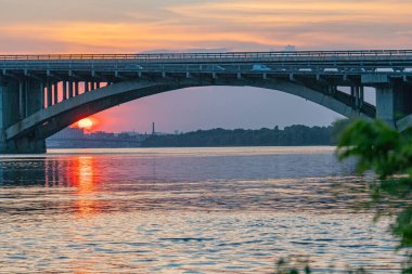 Görüntü, alacakaranlıkta bir su kütlesini kaplayan birden fazla kemerli sakin asma köprüyü gösteriyor. Yapı, kablolarla birbirine bağlı kuleler tarafından destekleniyor ve f Kyiv, Ukrayna 06-17-2024