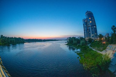 A panoramic photograph showcases modern urban waterfront architecture, featuring a striking tower with numerous balconies against a backdrop of setting sun The image highlig Kyiv, Ukraine 06-17-2024 clipart