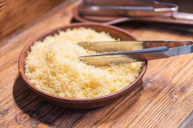 Ahşap kesim tahtasının yakın plan görüntüsü. Üzerinde iki spatulak maşası var. Kısmen rendelenmiş sarı parmesan peynirine batırılmış.