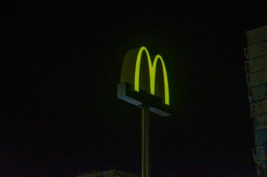 Parlak ışıklandırılmış bir McDonalds işareti karanlık bir zemin üzerinde duruyor, muhtemelen gece veya karanlık bir çevreyi işaret ediyor. Bu konum Kyiv, Ukrayna 08-24-2024