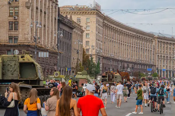Görüntü, Sovyet dönemi mimarisine sahip kentsel bir yapıyı, ana savaş tankı ve zırhlı personel taşıyıcıları gibi askeri araçların etrafında toplanan bir kalabalığı, muhtemelen ev Kyiv, Ukrayna 08-24-2024
