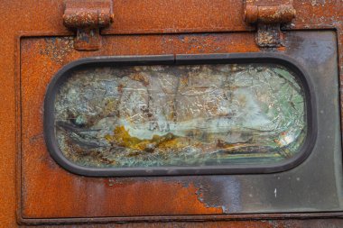 Görüntü, eski ve yıpranmış bir objeyi tasvir ediyor. Pencereye benzeyen paslı bir yapıya sahip. İçindeki cam gibi camlar toprak ve kirle gizlenmiş.