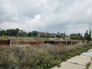 Zincirleme çitli bir açık hava sahnesi, arkasında aşırı gelişmiş ve kurumuş bitki örtüsü, arka planda düşük katlı metal yapılar, parçalı bulutlu gökyüzü, muhtemelen Kostiantynivka, Ukrayna, 06-15-2013