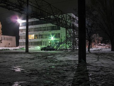 Kar ortasında ışıklı pencereleri olan bir sanayi binasının gece manzarası, ön planda kısmen donmuş su kütlesi, sol yapıda vinç, loş ışık ve gizli pho Kostiantynivka, Ukrayna, 01-15-2013