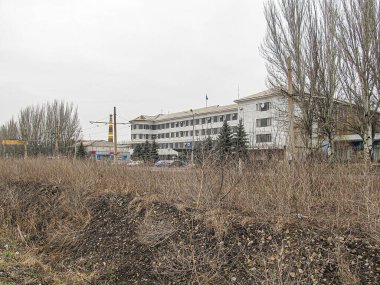 Görüntü, kuru, kahverengi ağaçlar ve seyrek bitki örtüsüyle çevrili beyaz, çok katlı bir bina, muhtemelen konut veya kurumsal bir manzara gösteriyor. Gr Kostiantynivka, Ukrayna, 01-15-2013