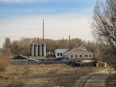 Gri silindirik tankların ve eğimli kiremit çatılı taş bir binanın egemen olduğu sanayi bölgesinde çimenler, ağaçlar, çalılar, bacalar, Kostiantynivka, Ukrayna, 01-15-2013
