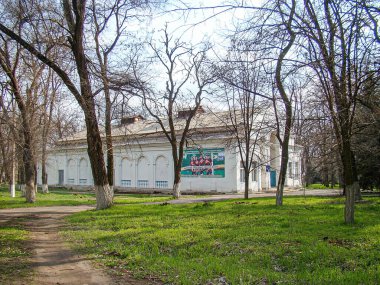 Simetrik kemer ve kolonlara sahip neoklasik ya da barok bir bina bir şehir parkındaki yaprak döken ağaçların arasında düz çatı, bulutlu gökyüzü ve yumuşak gölgeler arasında yer alır.