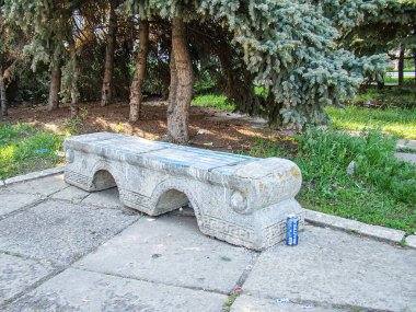 Görüntü, muhtemelen bir parkta ya da halk bahçesinde yıpranmış bir taş bankın bulunduğu açık hava sahnesini tasvir ediyor. Bankın etrafı yeşil yapraklı ağaçlarla çevrili. Bu da Kostiantynivka, Ukrayna, 06-15-2013
