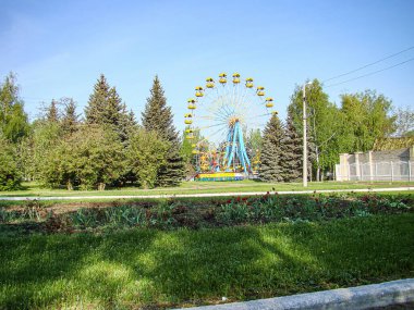 Canlı bir dönme dolap, yemyeşil bir alanda duruyor, yemyeşil ağaçlar, çimenler, toprak yamalar ve ona uzanan asfalt bir yol açık gökyüzü, Kostiantynivka, Ukrayna, 06-15-2013