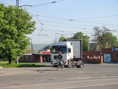 Beyaz bir teslimat kamyonunun arkasında bisiklet süren bir kişinin, kablolar, bitki örtüsü ve kırmızı tuğlalı bir bina, Kostiantynivka, Ukrayna, 06-15-2013