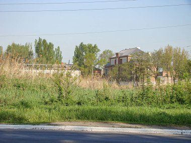 Görüntü, banliyöde yemyeşil bir aşırı büyümenin ortasında yıkık dökük, boş bir tuğla evi gösteriyor. Yakınlarda büyük bir ağaç duruyor ve açık mavi gökyüzü Kostiantynivka, Ukrayna, 06-15-2013