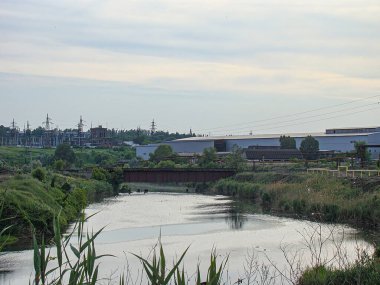 Bitkilerin, evlerin ya da binaların olduğu sakin bir nehir manzarası ve bulutlu bir gökyüzü altında maviden daha sıcak tonlara geçiş yapan bir köprü. Görüntü Kostiantynivka, Ukrayna, 06-15-2013 ile doğaldır.