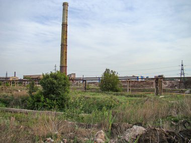 Parçalı bulutlu bir gökyüzünün altında terk edilmiş bir sanayi bölgesi, aşırı gelişmiş bitki örtüsü, enkaz ve fabrika yapılarından arta kalan uzun beton bacalar, Kostiantynivka, Ukrayna, 06-15-2013