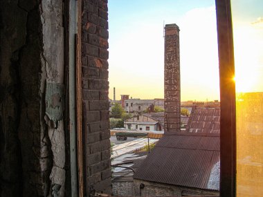 Görüntü, yıpranmış tuğlalar, soyulmuş boyalar ve hasarlı bir pencere çerçevesi ile eski bir binanın içini gösteriyor. Görüntü diğer binalara, temiz gökyüzü altındaki bacalara, Kostiantynivka, Ukrayna, 06-15-2013